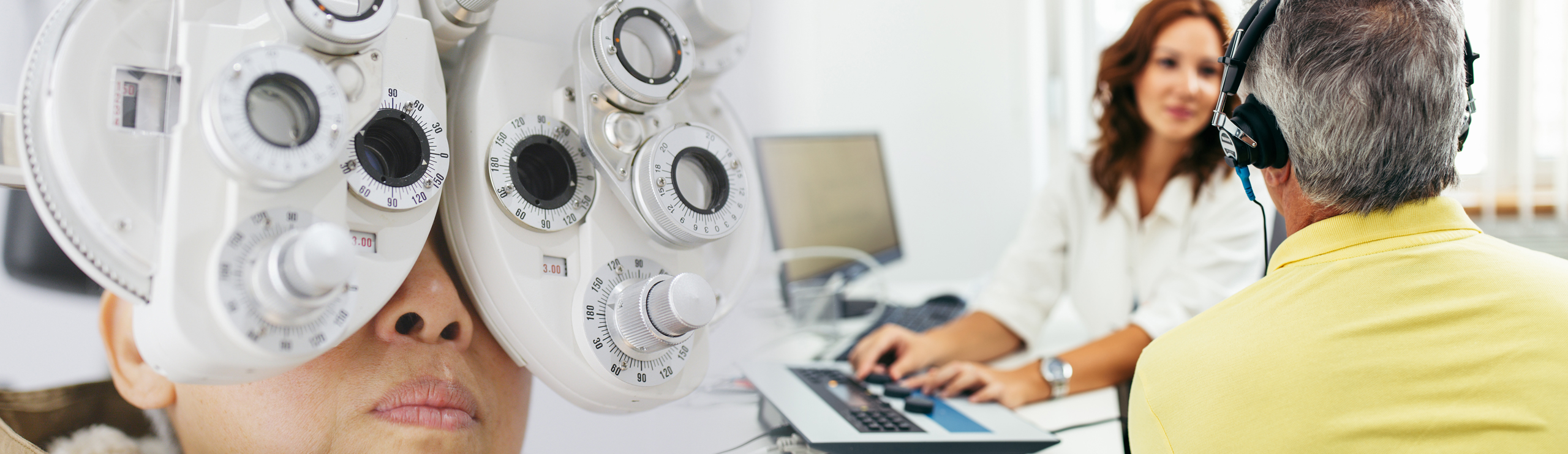 Patientin bei einer Augenuntersuchung mit einem Phoropter, im Hintergrund sitzt eine weibliche Ärztin am Computer und ein älterer Mann trägt einen Kopfhörer für einen Hörtest
