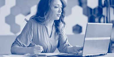 Frau arbeitet konzentriert am Laptop und macht Notizen mit Stift auf Papier