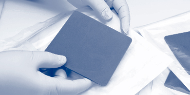 Hände mit Handschuhen halten eine quadratische, blaue Wundauflage
