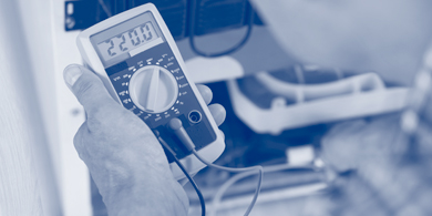 Hand hält digitales Multimeter mit Anzeige '220.0' vor technischem Gerät mit Kabeln