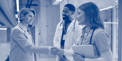 Zwei Personen geben sich die Hand, eine trägt einen weißen Arztkittel, die andere einen hellen Blazer, im Hintergrund steht eine weitere Person in Arztkittel