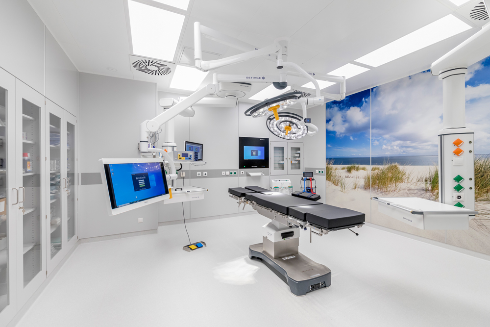 Moderner Operationssaal mit OP-Tisch, großen Monitoren, Decken-Operationsleuchten und Wandbild einer Strandlandschaft