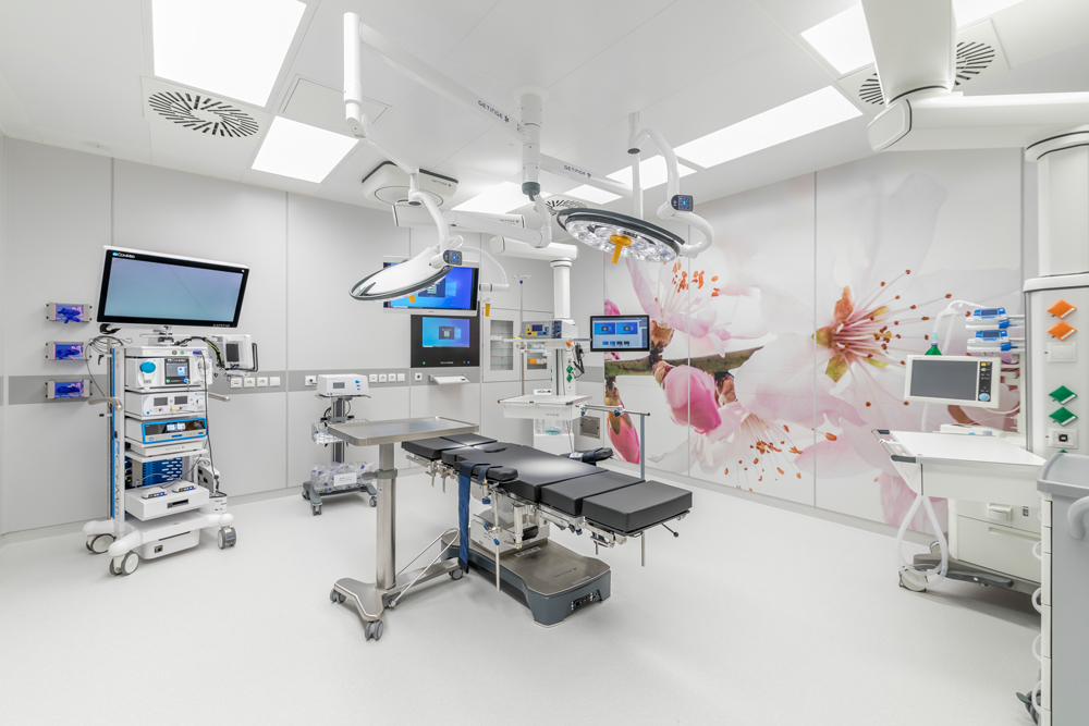 Moderner Operationssaal mit OP-Tisch, mehreren Monitoren, medizinischen Geräten und großer Wanddekoration mit rosa und weißen Kirschblüten