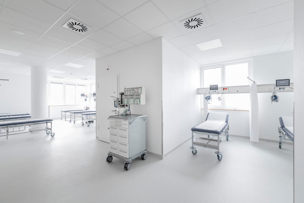 Helle, weiße Krankenhausstation mit mehreren leeren Patientenliegen auf Rollen und medizinischem Wagen in der Mitte