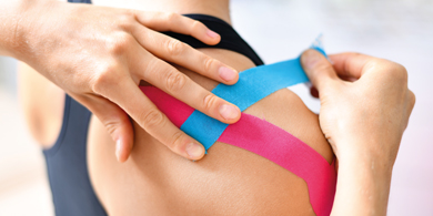 Person klebt einer Patientin einen blauen Streifen Kinesiontape auf die Schulter